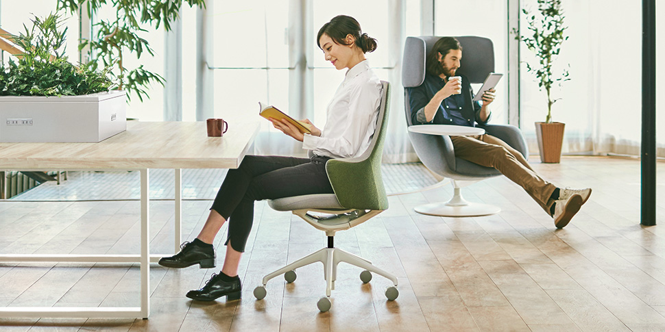 Muebles para oficina en un espacio de trabajo amplio y moderno, con una mujer leyendo en una silla frente a un escritorio y atrás un hombre en una silla tomando café relajándose.